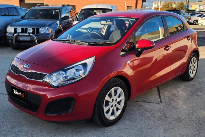 Front three-quarter view of red Kia Rio 2014 1.2 petrol manual rental car parked in front of apartment buildings in Tirana
